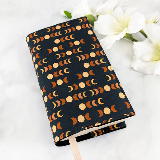 small adjustable fabric book cover, fabric showcases moon phases in beige and orange shades on a dark navy background.