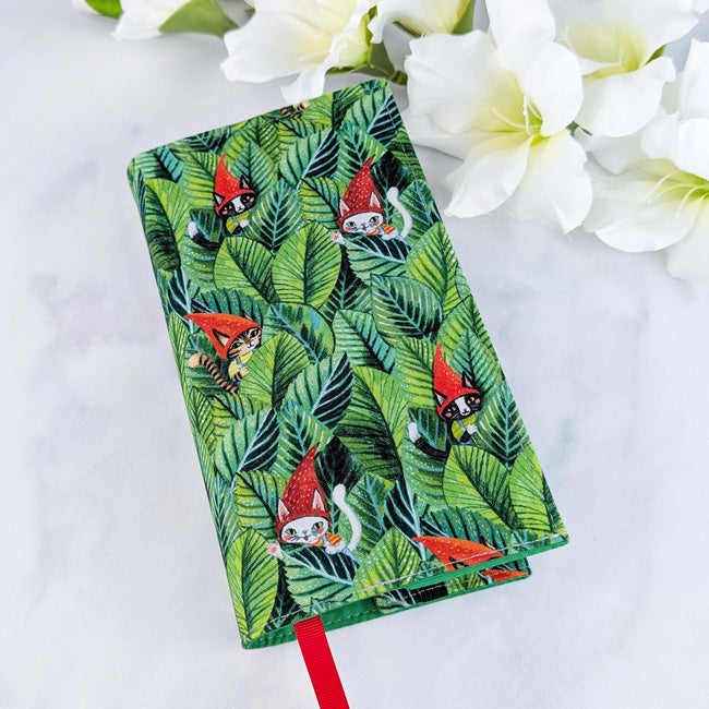 Fabric book cover featuring a pattern of cats wearing red gnome hats hidden among green jungle leaves, fitted on a book with a red ribbon bookmark, displayed beside white flowers.