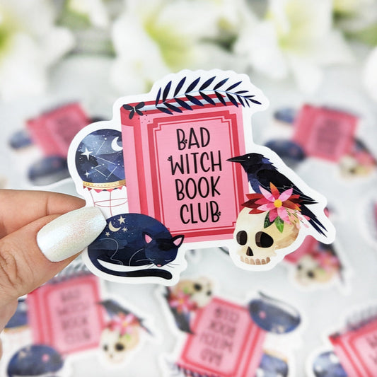 Vinyl sticker with the text “Bad Witch Book Club” on a pink book, decorated with a black cat, crow on a skull with flowers, and a crystal ball, held by a hand with iridescent white nail polish.