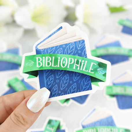 Vinyl sticker of a blue book with a green ribbon reading “Bibliophile,” held in a hand with white nail polish.