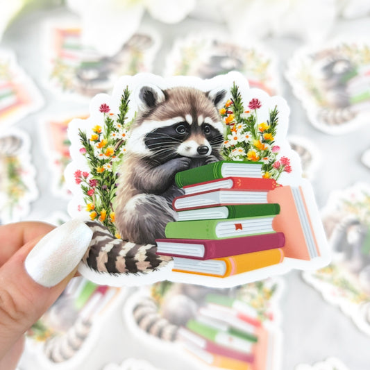 Illustrated sticker featuring raccoon with a stack of books and colorful flowers.