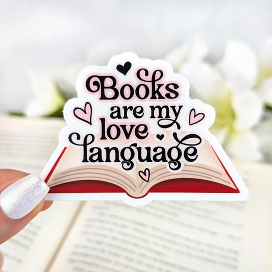 Vinyl sticker with an open book illustration and the phrase “Books are my love language” in decorative black and pink lettering with heart accents. Held by a hand with pearly white nail polish against a blurred book background.
