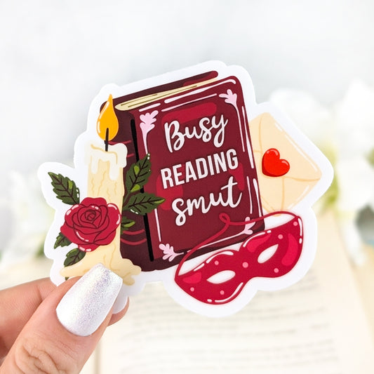 Romance reader vinyl sticker with book titled “Busy Reading Smut,” red mask, candle, rose, and love letter, held in hand over an open book background.