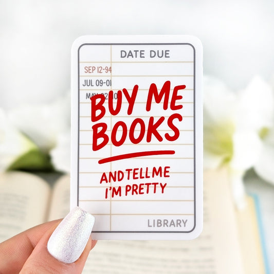 Vinyl sticker designed like a vintage library due date card with bold red text “Buy Me Books and Tell Me I’m Pretty,” held in hand over an open book.