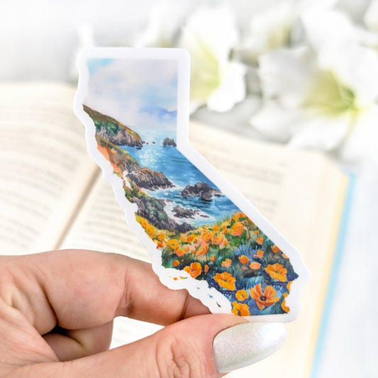 Vinyl sticker shaped like the state of California, featuring an illustrated coastal landscape with cliffs, ocean waves, and bright orange California poppies in the foreground. Held by a hand with iridescent white nail polish against a blurred book background.