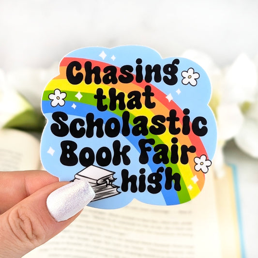 Sky-blue vinyl sticker with rainbow and stacked books reading “Chasing That Scholastic Book Fair High,” held in hand over an open book background.