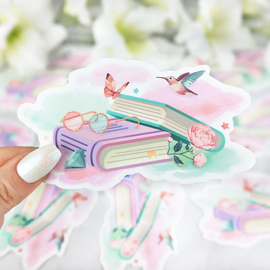 Vinyl sticker with pastel-colored books surrounded by a butterfly, hummingbird, pink roses, and glasses, set against a dreamy watercolor cloud background, held by a hand with iridescent white nail polish.