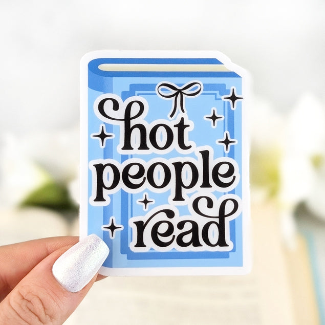 Blue book-shaped vinyl sticker with bold text “Hot People Read,” held in hand over an open book background with soft white flowers.