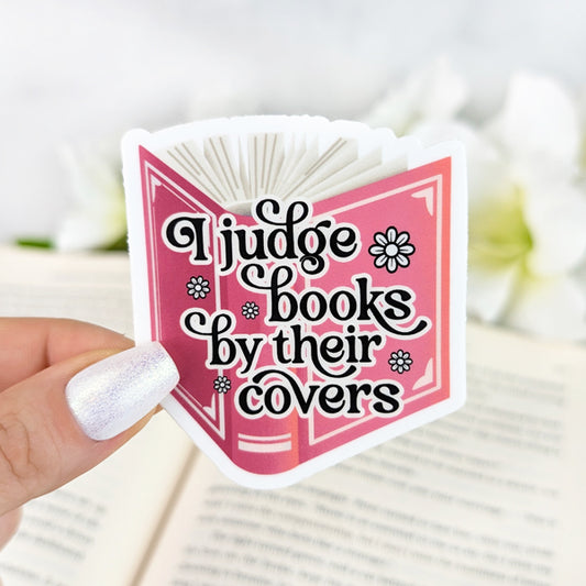 Vinyl sticker with the phrase “I judge books by their covers,” designed inside an open pink book with floral accents, held by a hand with shimmery white nail polish.