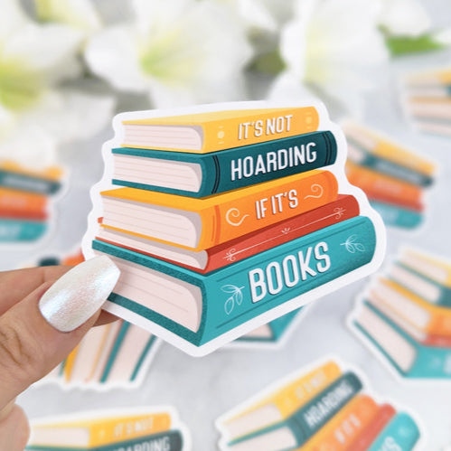 A vinyl sticker of a colorful stack of books with the text "It's Not Hoarding If It's Books," held by a hand with light nail polish. More identical stickers are scattered in the background.