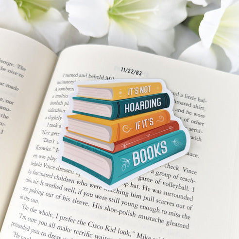 A vinyl sticker of a colorful stack of books with the text "It's Not Hoarding If It's Books," laying on a page of an open book.