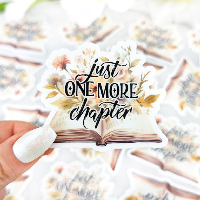 Watercolor sticker with a bunch of pastel flowers, open book and the text "Just One More Chapter" written in elegant calligraphy.