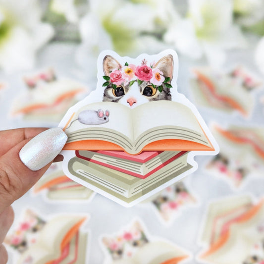 Vinyl sticker of a cat wearing a flower crown peeking over an open book with a tiny mouse illustration on the page, held by a hand with iridescent white nail polish.