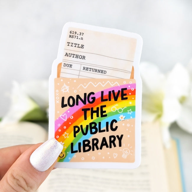 Vinyl sticker of a library card pocket with rainbow design and bold text “Long Live the Public Library,” shown held in hand over an open book.