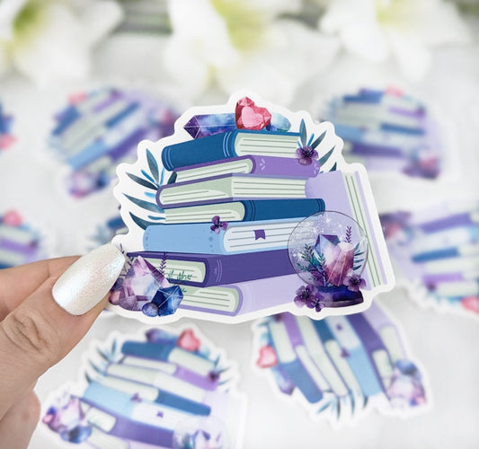 Vinyl sticker featuring a stack of purple and blue books decorated with crystals, gemstones, and flowers, held by a hand with iridescent white nail polish.