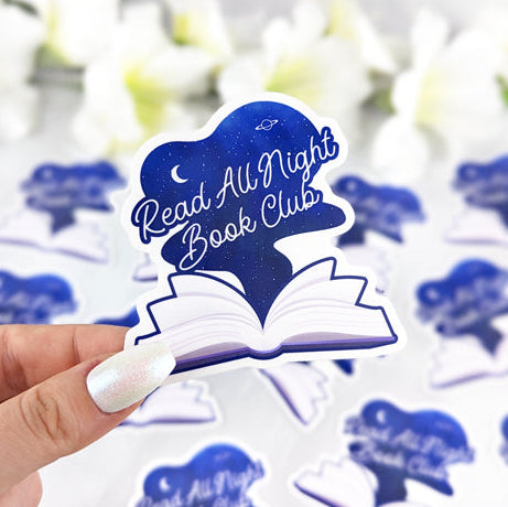 Vinyl sticker of an open book with a night sky and the text “Read All Night Book Club,” held by a hand with iridescent white nail polish.