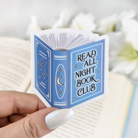 Illustrated sticker of a book with the text "Read All Night Book Club" in a vintage style held by a hand with light nail polish. Open book and white flowers in the background.