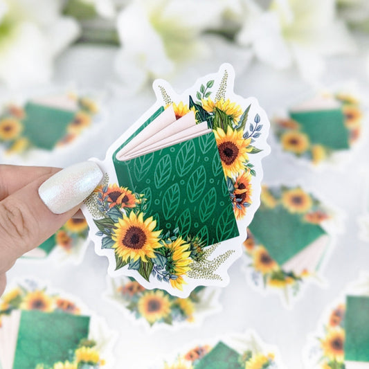 Vinyl sticker of an open green book decorated with sunflowers and leafy accents, held by a hand with iridescent white nail polish.