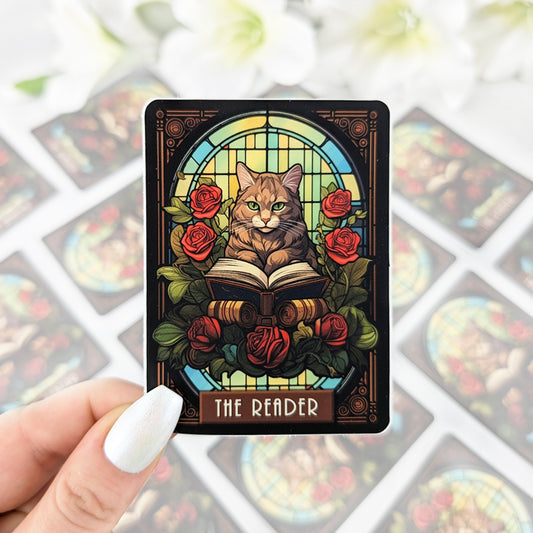 Vinyl sticker designed like a tarot card, featuring a cat sitting with an open book surrounded by red roses and stained glass, labeled “The Reader,” held by a hand with iridescent white nail polish.