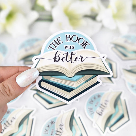 Illustrated sticker featuring a stack of books with the text "The Book Was Better" held by a hand with light nail polish. More identical stickers are scattered in the background.