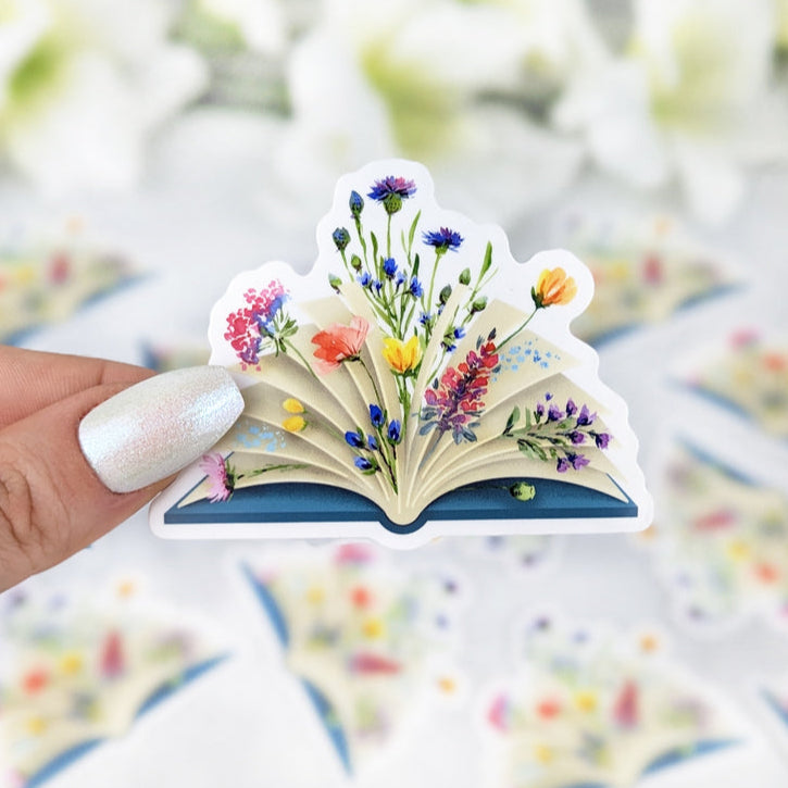 Illustrated sticker of a book overflowing with colorful wildflowers held by a hand with light nail polish. More identical stickers are scattered in the background.