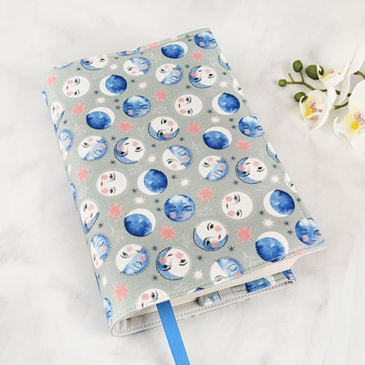 Fabric book cover with a whimsical celestial design featuring watercolor moon phases with smiling faces in shades of blue, white, and blush, scattered among soft pink stars on a muted gray background, finished with a bright blue ribbon bookmark.